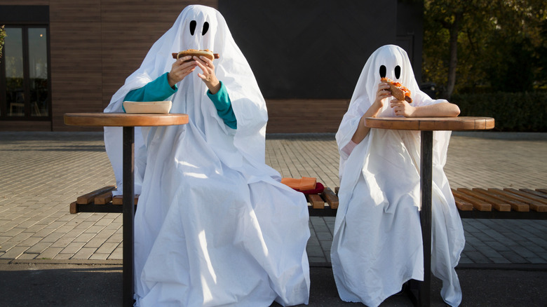 Two people dressed in sheep ghost costumes eating hot dogs at a table outside