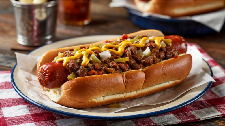A hot dog with fixings on top on a blue-rimmed plate and checkered napkin