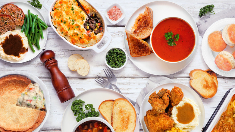 A table covered in plates of comfort foods like meatloaf and mashed potatoes with green beans, a shepherd's pie, grilled cheese with soup, fried chicken and mashed potatoes, chicken pot pie, and more