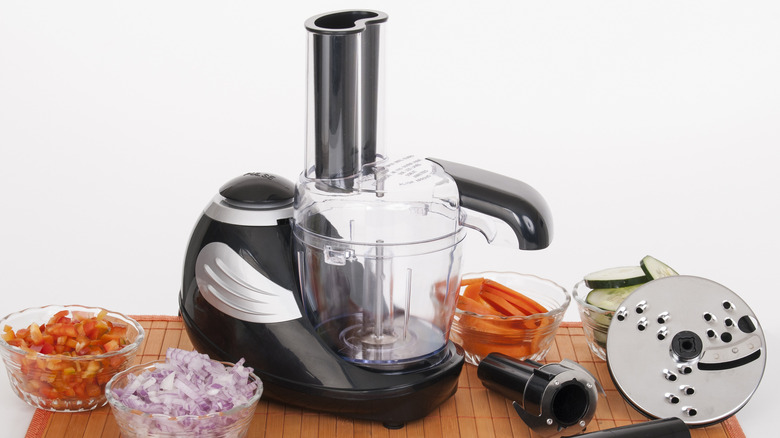 A food processor with accessories and bowls of chopped food on a wooden surface.