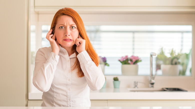 A woman with red hair and a white shirt in a kitchen covering her ears