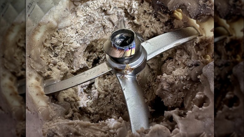 The detached blade from a Ninja Creami ice cream maker, sitting atop some partially churned ice cream, showing damage and metal stress where it broke off
