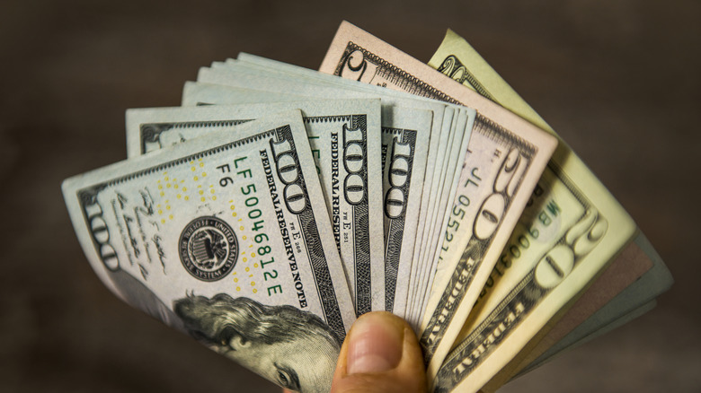 A hand holding a folded stack of US currency including $100 bills, $50 bills, and $20 bills