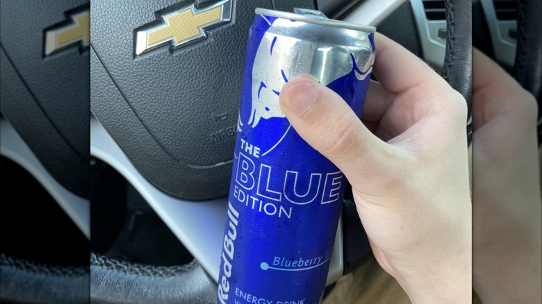 Hand holding open can of Blue Edition Red Bull in front of car steering wheel, mirrored image