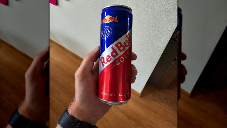 Mirrored image of hand holding can of Red Bull Cola in front of white wall
