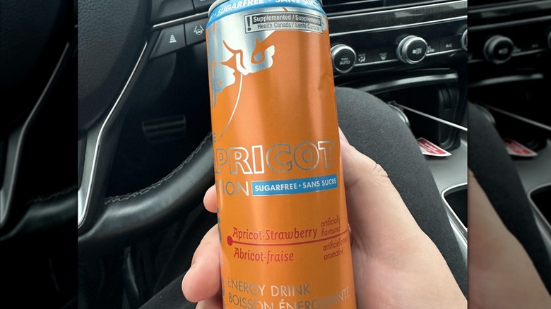 Hand holding Red Bull's Sugarfree Strawberry Apricot Amber Edition in front of car steering wheel, mirrored image