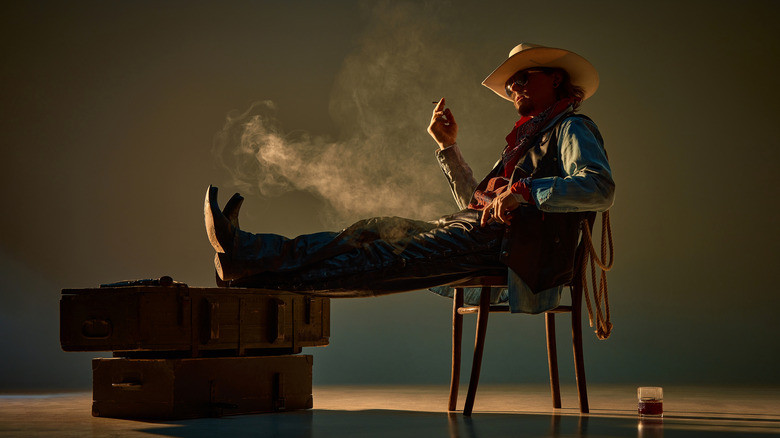 A stylizes shot of a cowboy sitting on a chair with glass of liquor on the floor