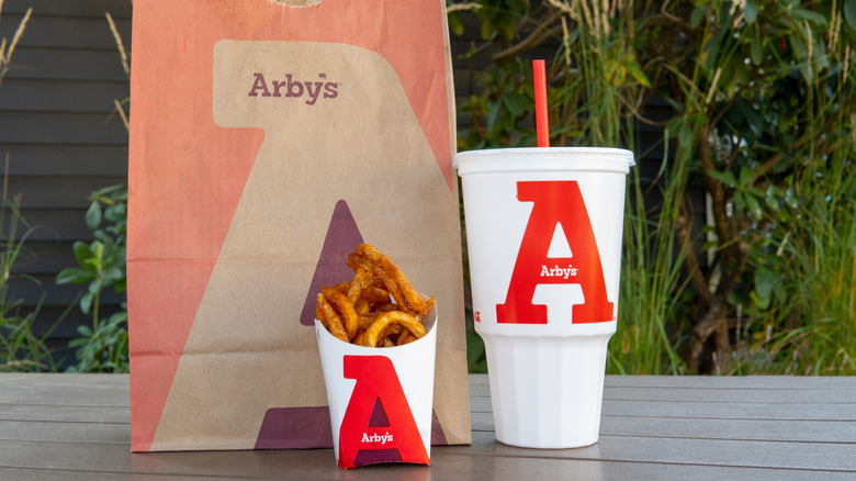 Arby's curly fries and drink in front of a bag from the chain on a table.