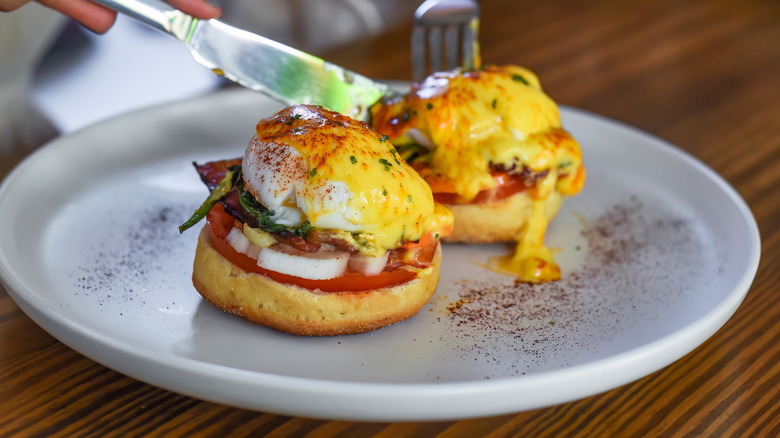 Someone cuts into two pieces of eggs Benedict topped with herbs and paprika