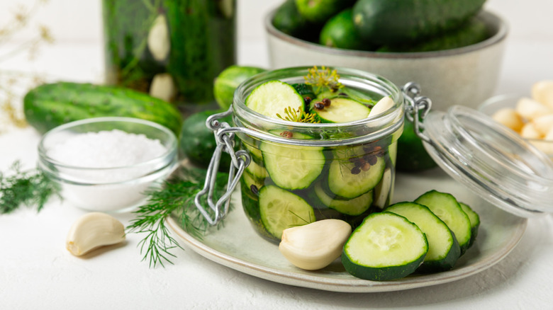 Small glass container of sliced homemade pickles on silver dish next to sliced pickles, bowl of salt, garlic, and fresh cucumbers
