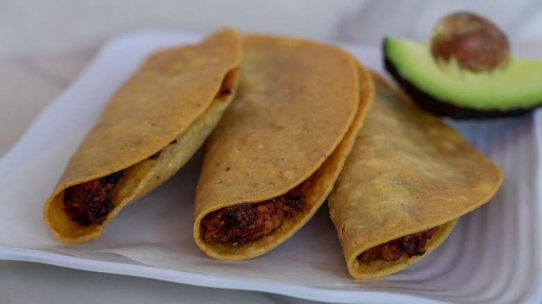 Fried tortilla tacos on a plate with part of an avocado