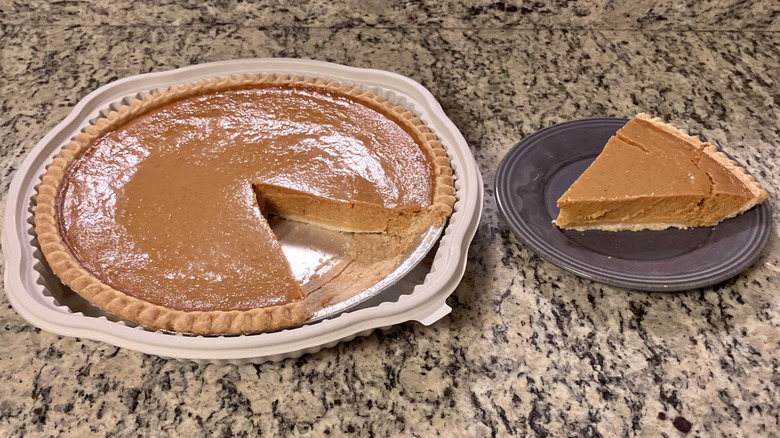 A whole Costco Kirkland Signature Pumpkin Pie sits next to a slice of the pie that's on a purple plate