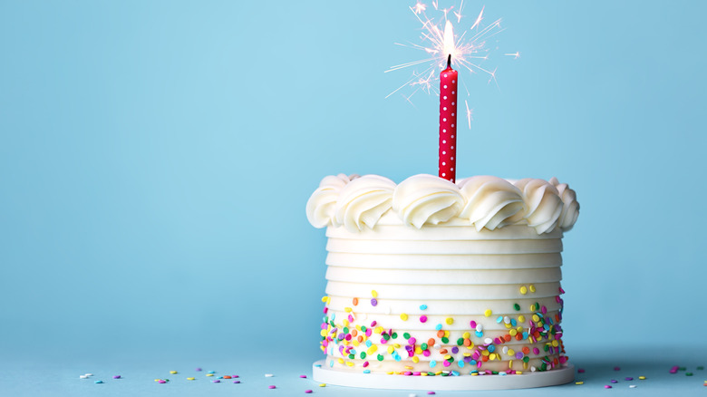 A small white circular birthday cake with a lit red candle against a blue studio background.