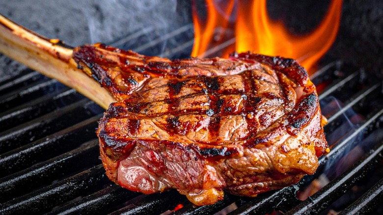 A Tomahawk Steak on the grill, covered in grill marks, with flames flaring up in the background