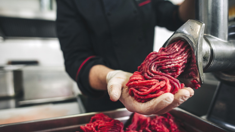 A gloved hand catching fresh ground beef coming out of a meat grinder.