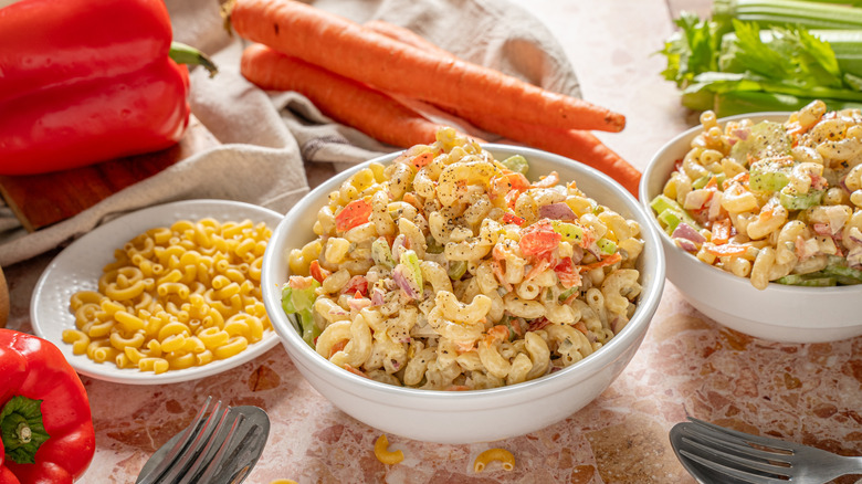 Two bowls of pasta salad on a table top next to a plate of plain pasta with vegetables in the background