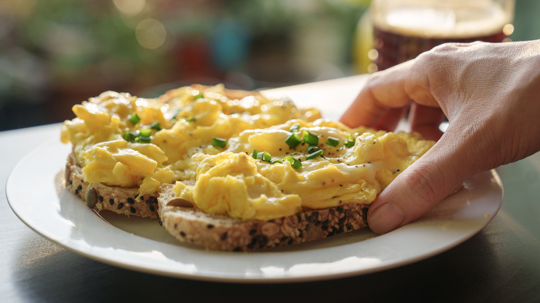 A hand reaches for a slice of bread topped with scrambled eggs and fresh herbs