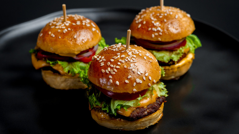 A trio sliders for sesame seed buns, lettuce, sauce and other veggies on a black plate