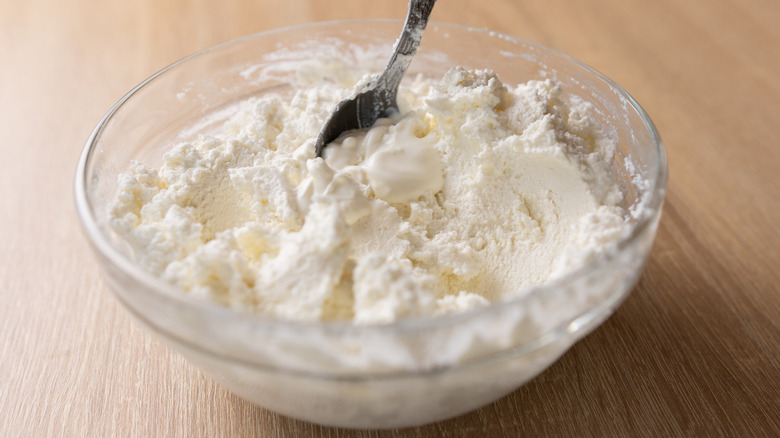 spoon mixing sour cream into crem cheese, shown in a glass mixing bowl on a wooden, Scandinavian-looking counter or tabletop