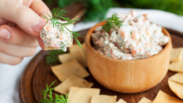 hand scooping smoked salmon and dill dip from a wooden bowl, on a wooden serving tray, with fresh dill garnish visible on the individual portion as well as the main bowl