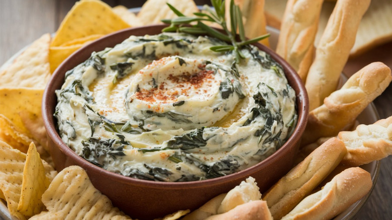 creamy spinach dip garnished with paprika and fresh rosemary, served in a brown bowl and surrounded with waffle-style crackers and crisp bread sticks