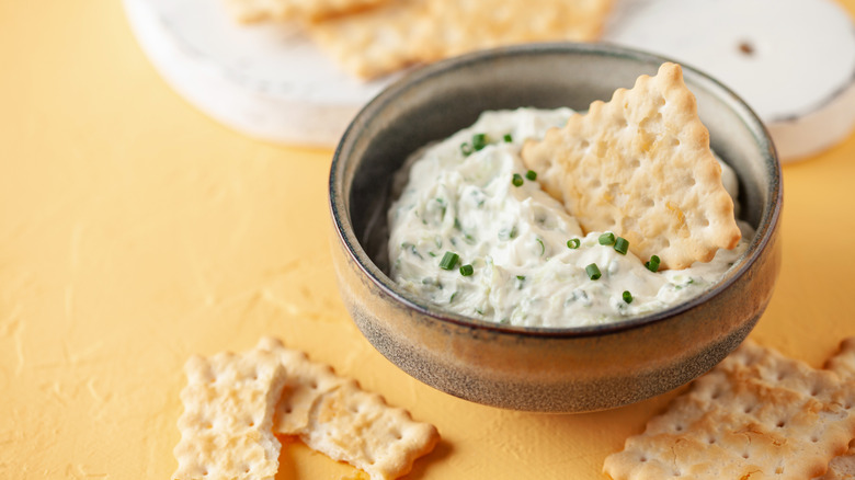 a bowl of creamy-looking dip with chopped chives, shown in a rustic brown bowl, surrounded by crackers, with one cracker in the dip and more visible behind the bowl, but out of focus