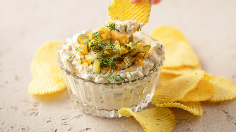 fried pickle dip, shown on a stone countertop with a pickle-and-dill garnish, surrounded by ripple potato chips, with a chip being dipped into the rear of the bowl
