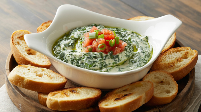 spinach dip in a white bowl, garnished with diced tomato and sliced green onion, served on a platter with toasted baguette slices