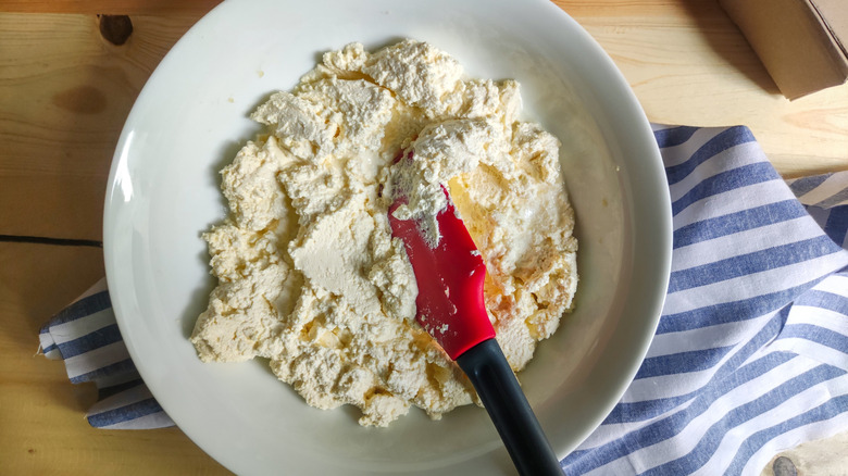 softened cream cheese shown in an overhead view, in a white mixing bowl with a black-and-red silicone spatula, resting on a blue and white striped kitchen towel and a pale wooden tabletop