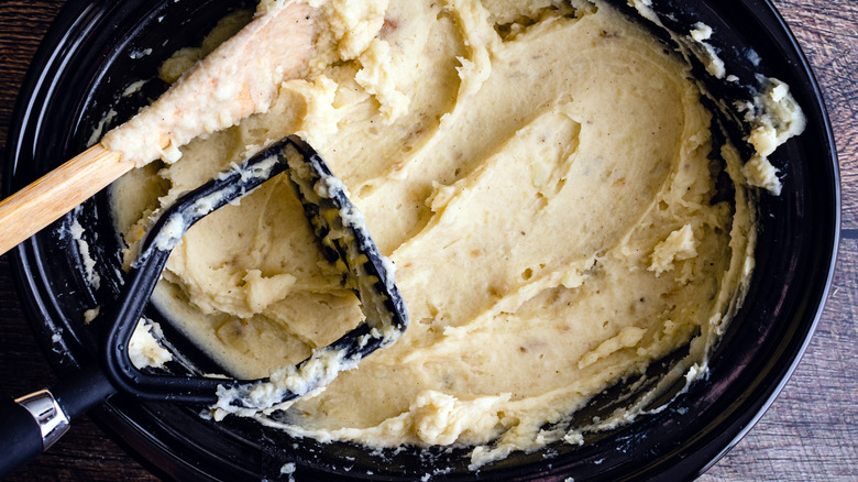 Slow cooker filled with mashed potatoes with wooden spoon and potato masher resting on top