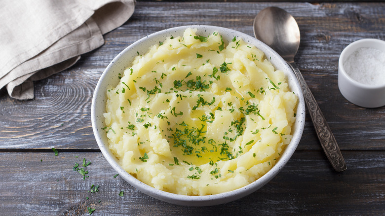 Small white bowl of mashed potatoes with butter and dried parsley with spoon and cloth napkin on wood surface.