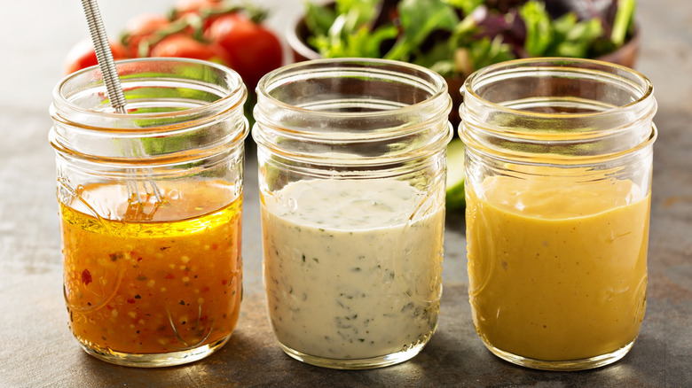 Three jars of different homemade salad dressings with out of focus salad and tomatoes in the background