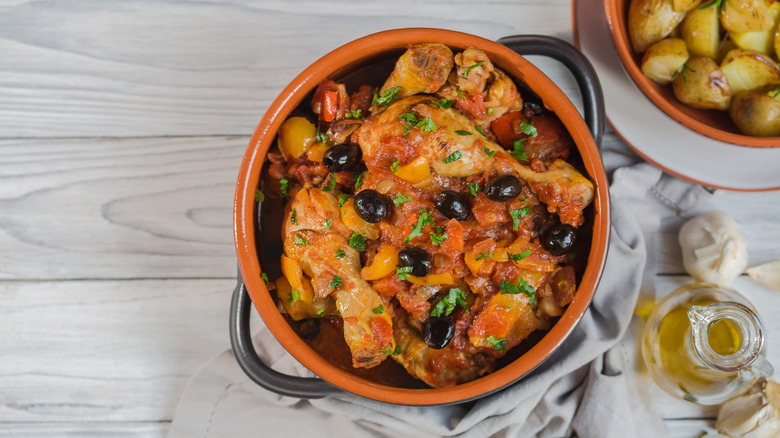 a pot of chicken cacciatore beside a bowl of potatoes, a bottle of olive oil, and a head of garlic
