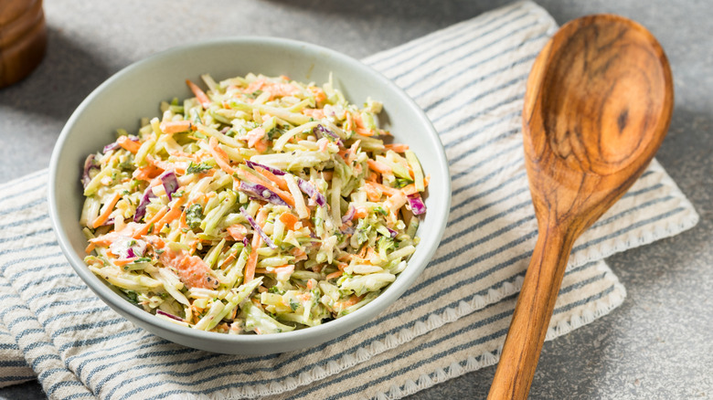 Pale green bowl of coleslaw sitting on striped kitchen towel next to wooden spoon