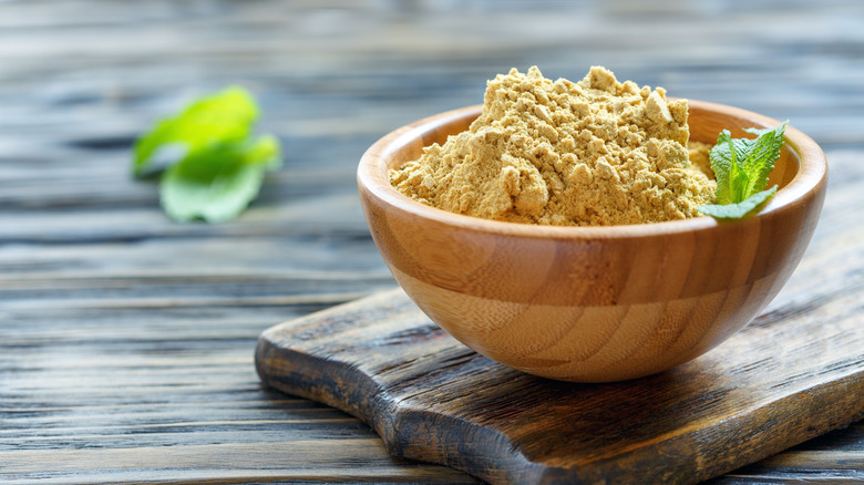 Dry mustard powder in light wooden bowl sitting on dark wooden board with green herb decoration