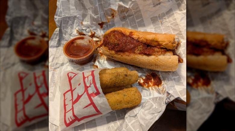 A Sheetz meatball sub with a container of marinara sauce beside Sheetz mozzarella sticks