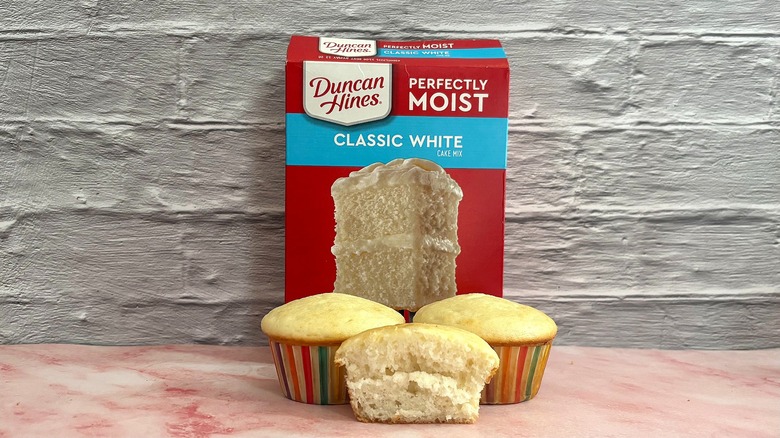 Box of Duncan Hines Classic White cake mix behind three cupcakes on grey background