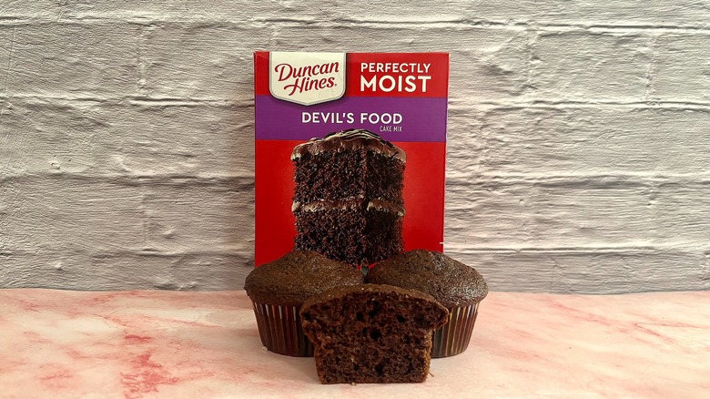 Box of Duncan Hines Devil's Food cake mix behind three cupcakes on grey background