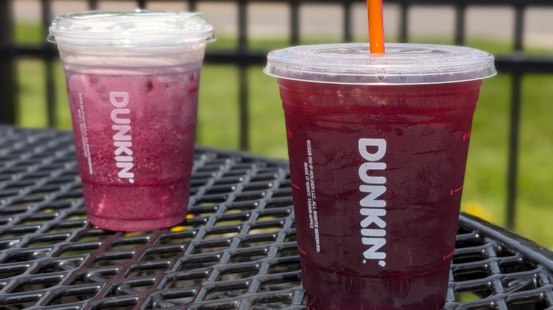A purple Dunkin' drink with a straw in it in front of a purple Dunkin' drink with oatmilk and cold foam