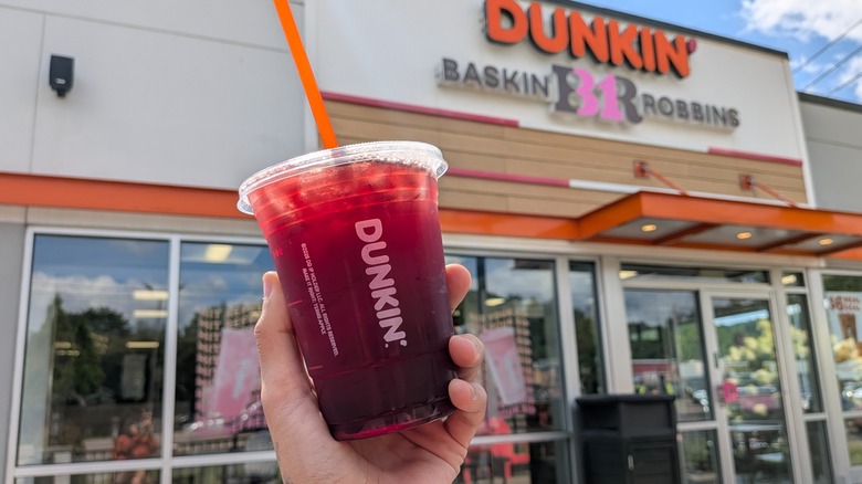 A hand holding a purple Dunkin' drink with a straw in it in front of a Dunkin'-Baskin Robbins storefront