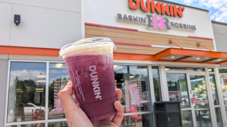 A hand holding a purple Dunkin' drink with cold foam in front of a Dunkin'-Baskin Robbins storefront