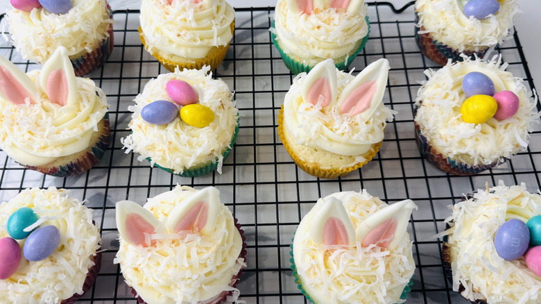 white-frosted cupcakes topped with shredded coconut and Easter-themed decorations sitting on a wire rack