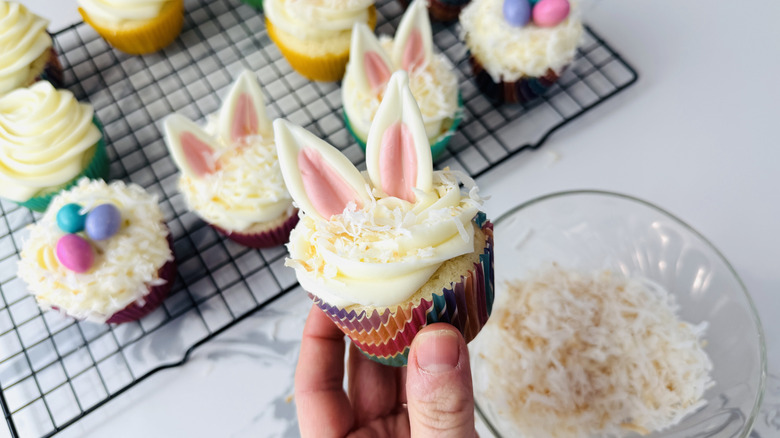 hand holding a white-frosted cupcake topped with coconut and a pair of pink and white pointed oval shapes