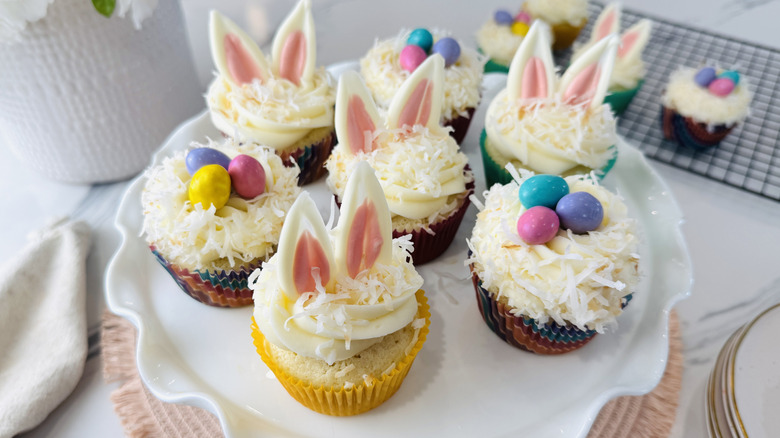 white-frosted cupcakes topped with shredded coconut and Easter-themed decorations sitting on a white plate