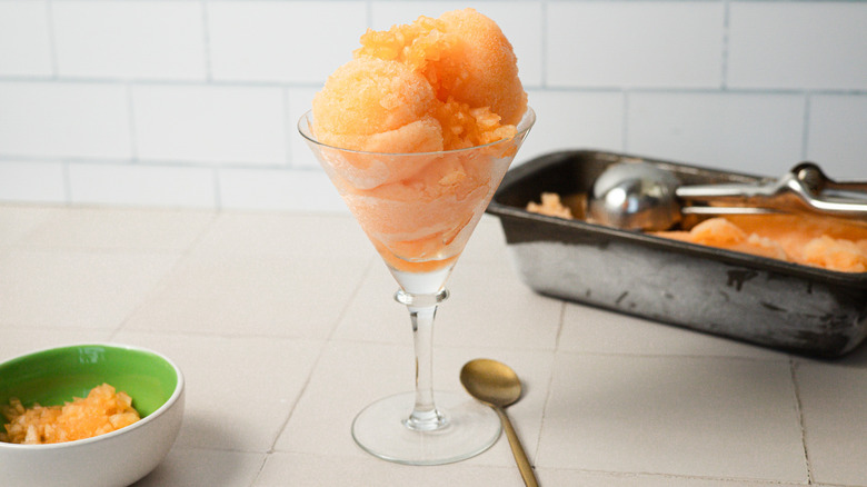 A glass of cantaloupe sorbet sits beside a spoon, a pinch bowl with diced cantaloupe and a container of ice cream in the back