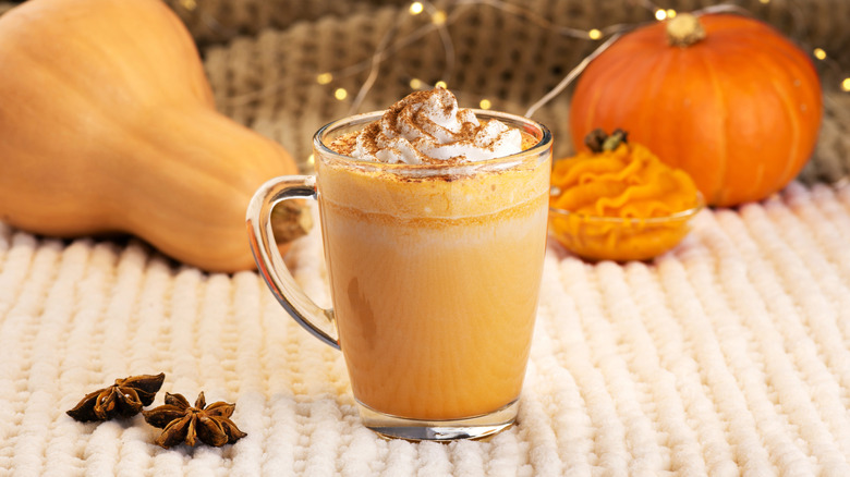 Pumpkin spice latte in clear glass mug with whipped cream and spice garnish with squash and pumpkin in background