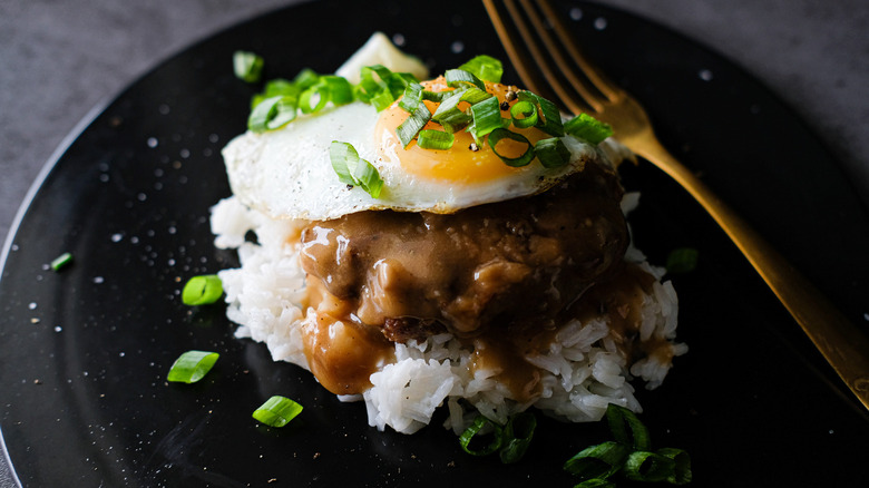 Loco moco garnished with scallions on a black plate with a gold fork