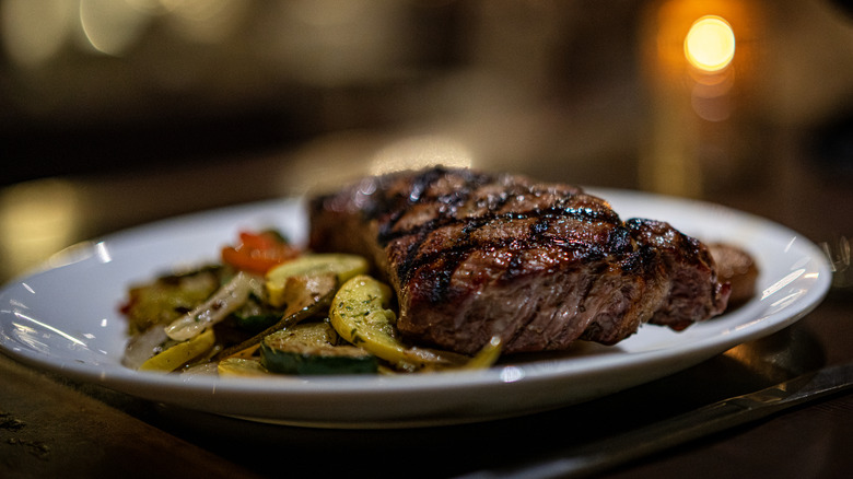 A plated steak accompanied by some cooked vegetables rests on a table beside a knife, with a blurred wine glass and candle in the background.