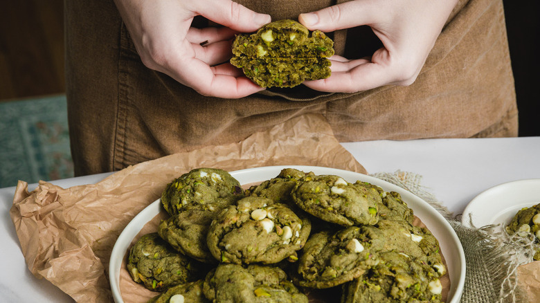 Hands holding one white chocolate matcha cookie, cut in half, over a large platter of cookies