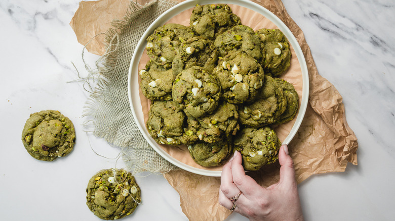 Hand taking one white chocolate matcha cookie from a white plate filled with cookies